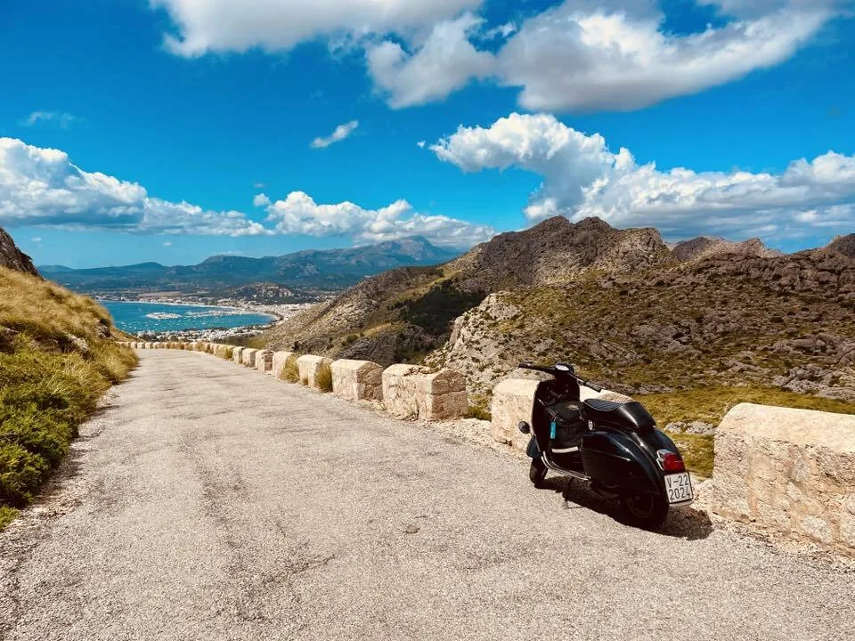 Valentina 1967 Vespa GS150 on the road to Cap Formentor Mallorca with bay and Tramuntana mountains in background — Vuelta Balearica 2026