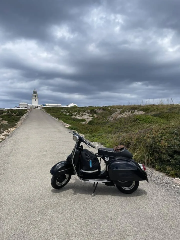 Valentina 1967 Vespa GS150 on the road to a Menorca lighthouse under dramatic cloudy sky — Vuelta Balearica 2026