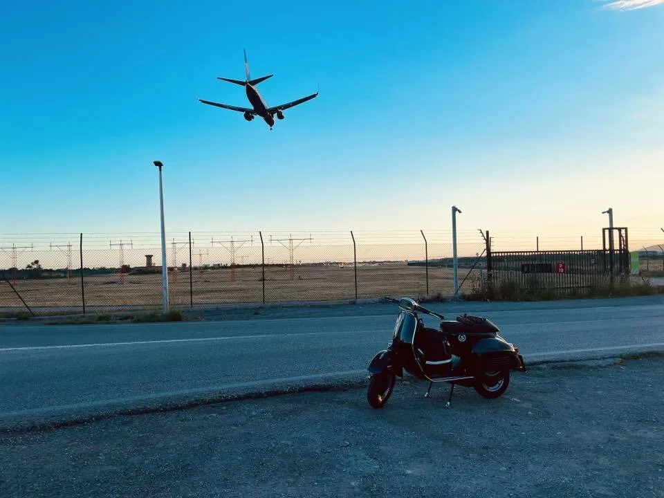 Valentina 1967 Vespa GS150 parked at Palma airport perimeter road at sunset with plane landing overhead — Vuelta Balearica Mallorca 2026