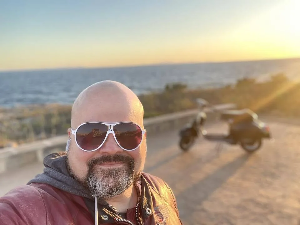 Markus André Mayer selfie at sunset on Menorca coast with Valentina Vespa GS150 in background — Vuelta Balearica 2026